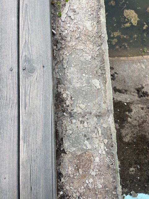 Gammal poolkant med borttagna stenplattor bredvid trätrall, poolen är tömd och ytan är ojämn och krackelerad, redo för renovering.