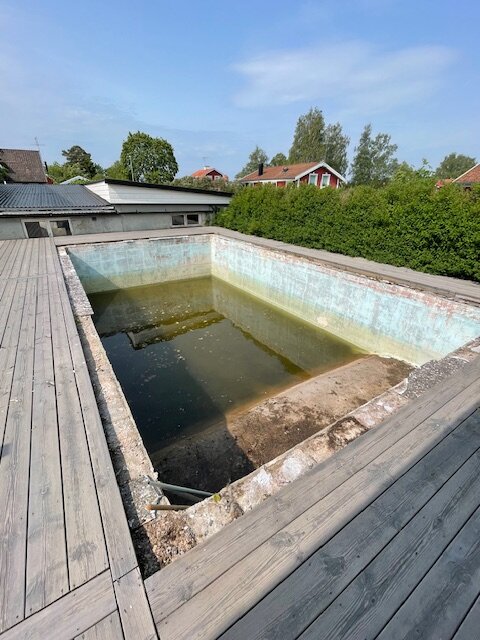 Tom, gammal pool med trasiga stenkanter, omgiven av ett trädäck. Poolens botten är delvis fylld med vatten och alger. Renoveringsbehov syns tydligt.