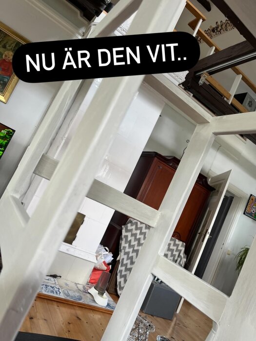 Vitmålad benställning för köksskåp i ett rum med trägolv, öppen spis och tavlor, texten "NU ÄR DEN VIT.." syns över bilden.