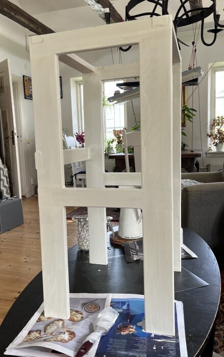 Vitmålad benställning för köksskåp står på ett bord, omgiven av tidningspapper och målarutrustning i ett vardagsrum.