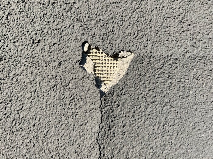 Skadad putsfasad med spricka där glasfiberremsa och underliggande material syns. Plastfärg har släppt från ytan.