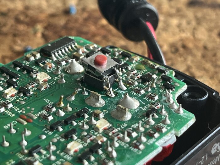 Närbild på kretskort med en röd knapp monterad på, omgiven av lödpunkter och elektroniska komponenter.
