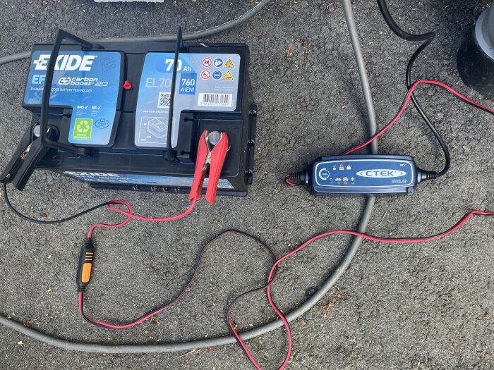 Bilbatteri med klämmor anslutna, kopplat till en CTEK-laddare på marken.