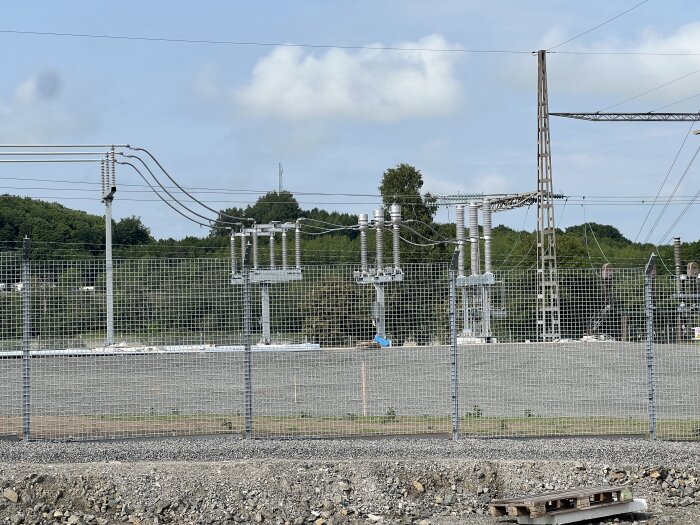 En transformatorstation med kraftledningar och ställverk, omgiven av ett stängsel, mot en bakgrund av träd och klar himmel.