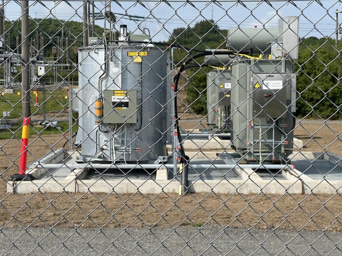 Transformatorstation med flera gråa och metalliska enheter omgärdade av ett nätstängsel, set på mark med vegetation och kraftledningar i bakgrunden.