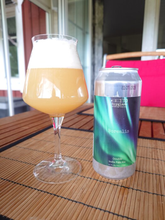 Ett glas med skummande DIPA från Spyglass Brewing Company bredvid en burk med etiketten "Borealis", placerad på ett bord utomhus.