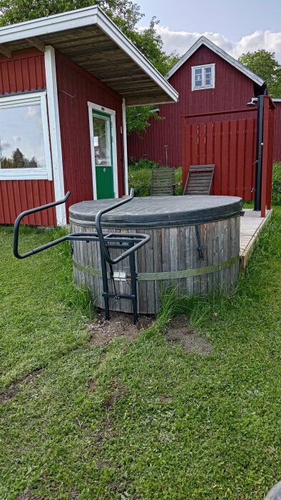 Badtunna med lock och stöd, omgiven av gräs, med en röd byggnad och uteplats med stolar i bakgrunden.