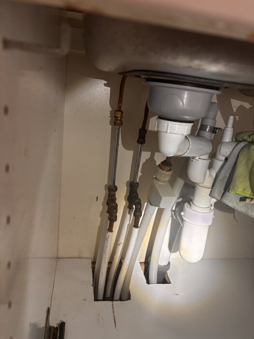 Rörsystem under ett handfat med flera vita plast- och metallrör, kopplingar och en avloppsdel i badrumsmiljö.