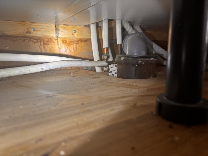 Rör och kopplingar under diskbänk, med vita och metalliska rör samt ett brunt järnliknande rör placerade mot en träbakgrund.