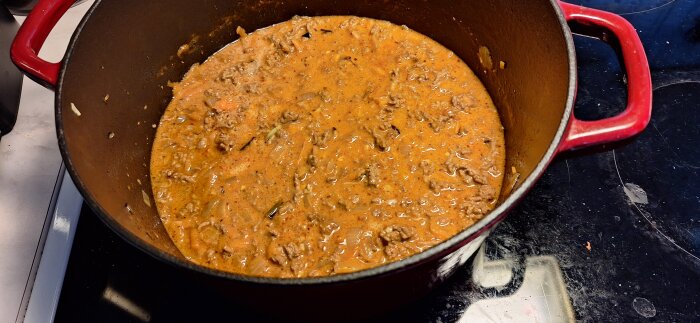Köttfärssås i en röd gryta på en spis, med synliga köttbitar och lök i såsen.