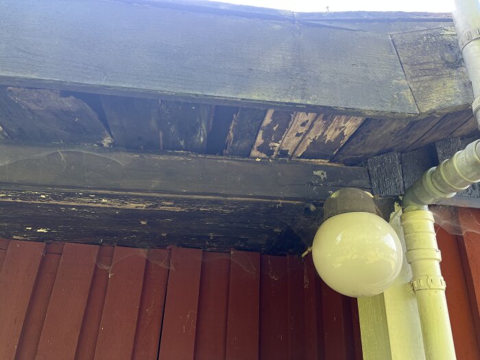 Skadat vindskydd och takdel på hus, träet är slitet och delvis ruttnat. Ser även en rund vit lampa och delar av ett stuprör vid väggen.