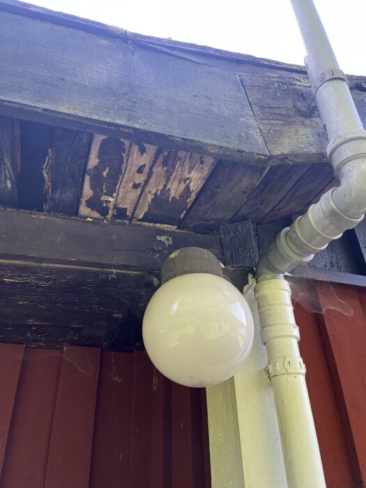 Skadat vindskydd med flagnande träpanel under plåttak, bredvid stuprör och lampa på ett hus.