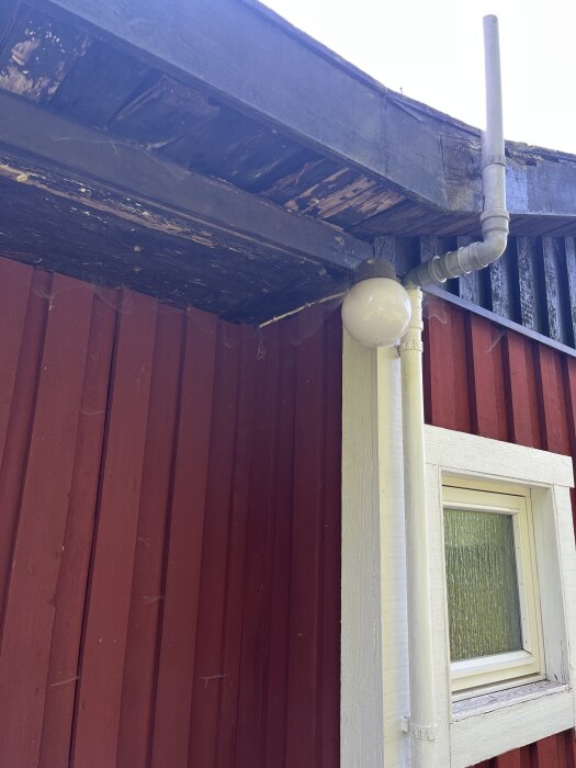 Rött hus med skadad vindskena där träet är slitet och delvis blottat; kompletteras av vitmålad takränna och takfot.