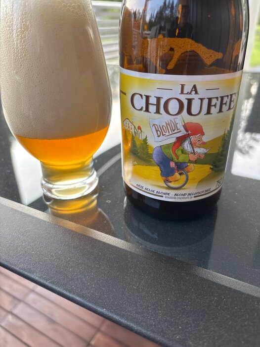 En stor flaska La Chouffe Blonde öl står bredvid ett glas med skummande öl på ett glasbord.