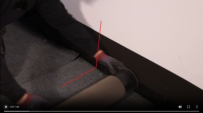 En person lägger takpapp längs en vägg och golvkant, med en rulle av takmaterial.