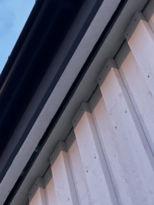 Vit takfot med smal profil och fasad. Blond trästruktur syns mot blå himmel i bakgrunden.