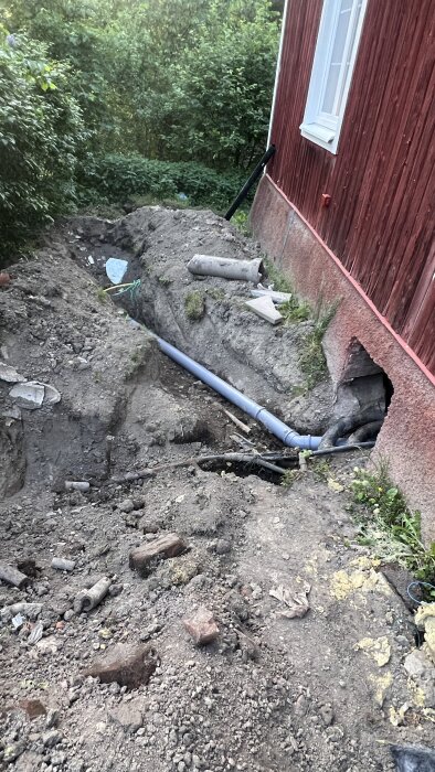 Grävd schakt med nyinstallerat avloppsrör av plast utanför ett rött hus. Mark är delvis uppgrävd med gamla rördelar synliga.