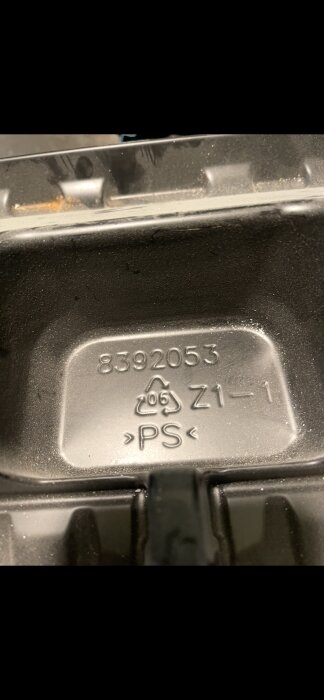 Text och symboler präglade på en svart plastdel, inklusive återvinningssymbol och texten "8392053 Z1–1 >PS<".