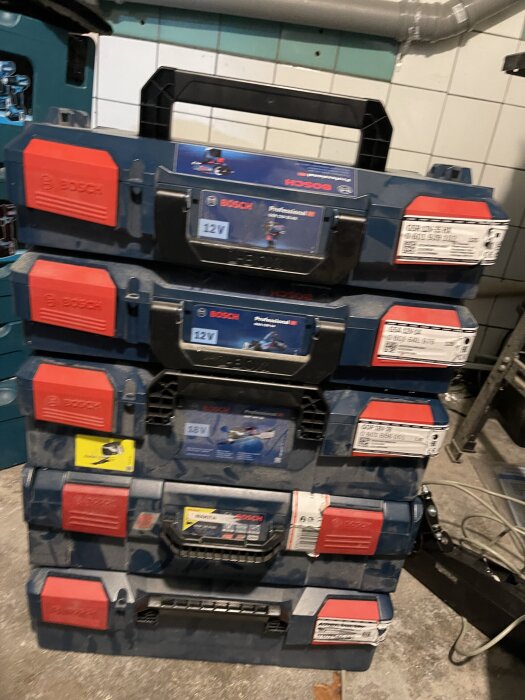 Staplade blå Bosch-verktygslådor med röda handtag, placerade på ett golv nära vit kakelvägg, text och etiketter synliga på lådorna.