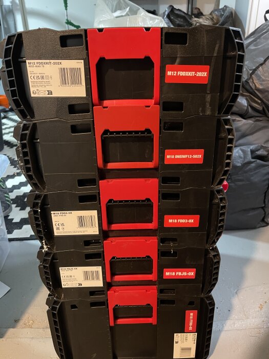 En stapel med svarta och röda verktygslådor med märken som M12 FDDXKIT-202X etiketterade på framsidan, på ett golv.