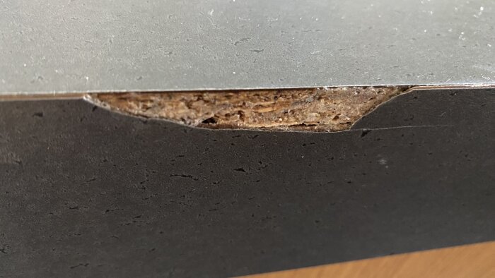 Närbild av en skadad yta på en möbel där laminatet har lossnat, vilket exponerar materialet under ytan.