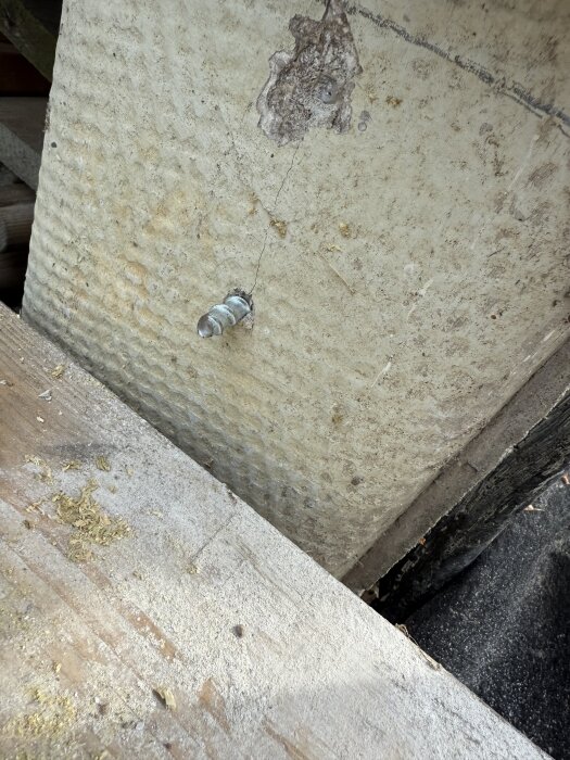 Betonipylvääseen kiinnitetty murtunut ruuvi puupinnan yläpuolella. Näkyvissä myös rakennuspölyä ja vaurioitunut kiinnityspinta.