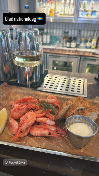 Räkor serverade på en bricka med citron, bröd och sås bredvid ett glas vitt vin, fotograferat på en bar.