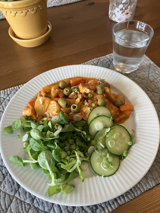 Tallrik med rotfruktsgratäng, toppad med oliver, bredvid sallad, gurkskivor och ett glas vatten.