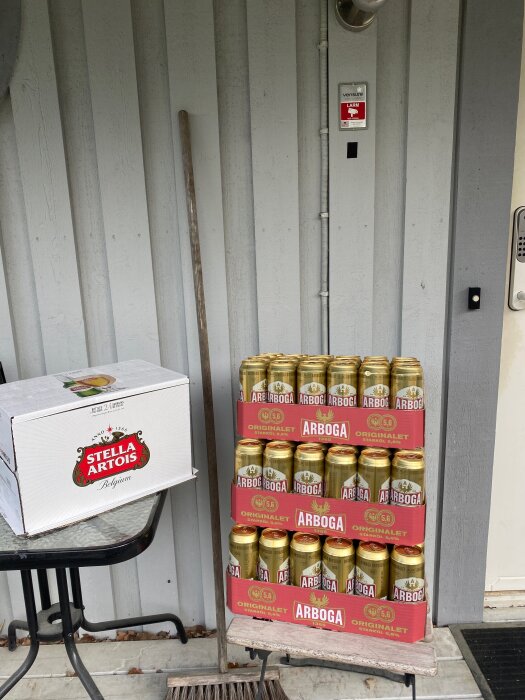 Låda med Stella Artois och staplade Arboga-ölburkar utanför en dörr med larmskylt.