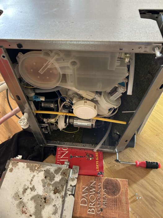Inre delen av en diskmaskin med synlig avloppsslang och andra komponenter. Verktyg och böcker syns på golvet framför maskinen.