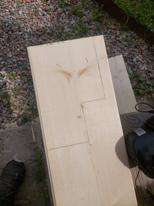 Närbild av en träskiva med märkningar för snitt, del av ett pågående byggprojekt på en grusig markyta.