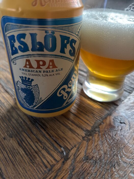 Burk och glas med Eslövs APA, en American Pale Ale, 5,2% alkohol, står på ett träbord med gyllene öl och skum i glaset.