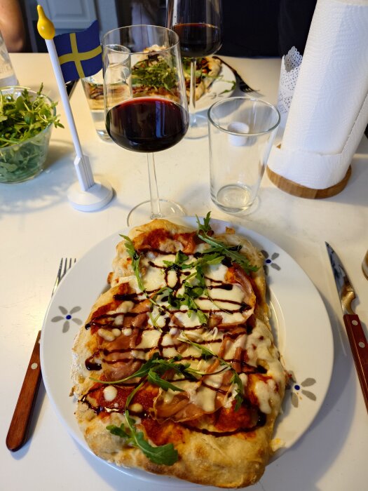 Pinsa med prosciutto, ruccola och balsamico på ett bord med vinglas, flagga och pappersrulle.