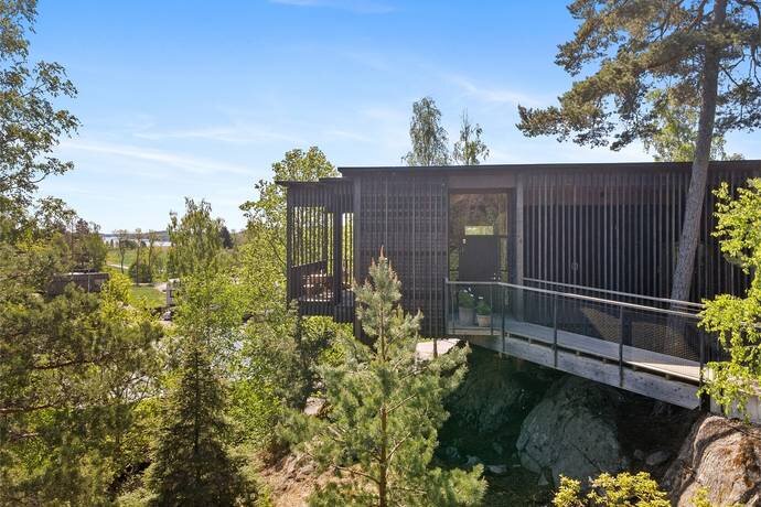 Modernt hus med träfasad i skogsmiljö, beläget på en klippa med utsikt över omgivande träd och klarblå himmel.