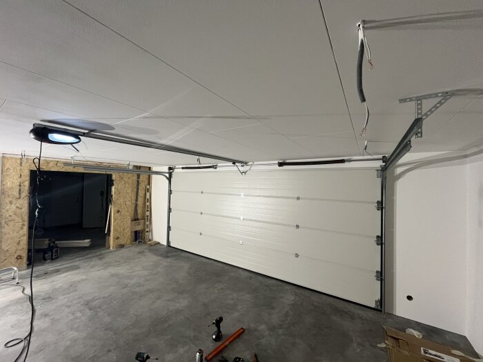 Inre vy av ett garage med en stor vit takskjutsport monterad, några verktyg och kartonger ligger på betonggolvet.