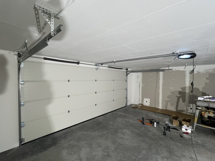 Garage med en nyinstallerad, inåtgående takskjutsport och verktyg på golvet, betonggolv och delvis uppsatta väggpaneler.