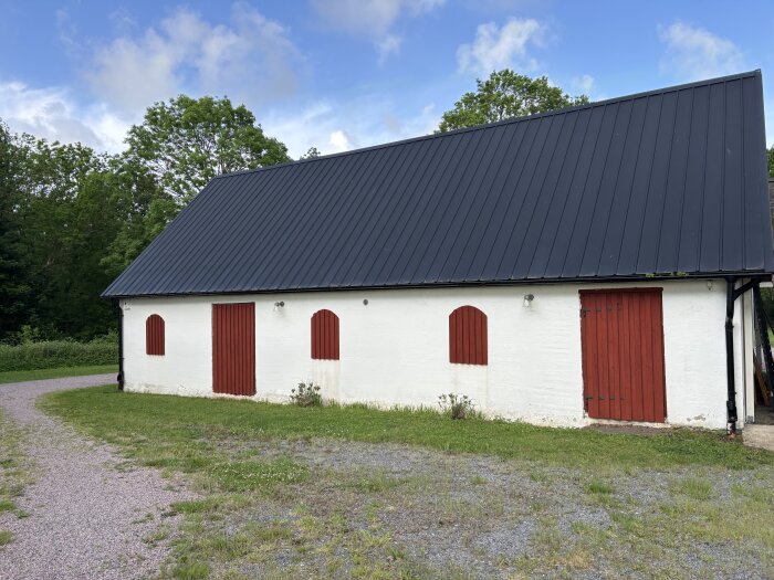 Vit lada med röda dörrar och skivor, plats för fönster. Grönt gräs och grusväg i förgrunden, trädtäckta kullar i bakgrunden.
