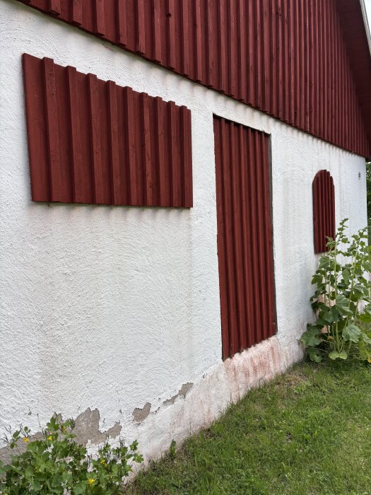 Rödvit lada med täckta fönsteröppningar och sliten fasad, omgärdad av grönska.