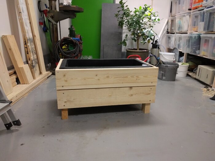 Träplanteringslåda med murarbalja inuti, placerad i en verkstad med byggmaterial och en krukväxt i bakgrunden.