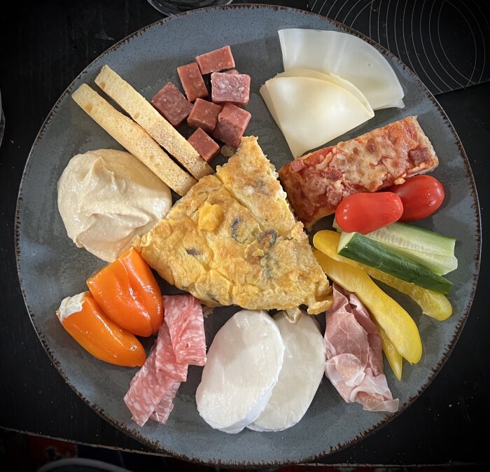 En tapastallrik med spanska ostar, charkuterier, omelett, grönsaker, bröd och krämig hummus.