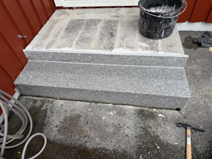 Steg av grå granit framför en röd byggnad med en hink på och en hammare vid sidan.