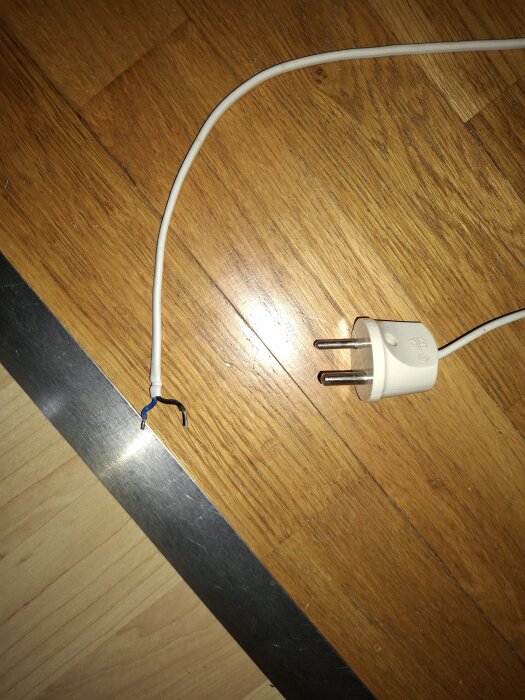Vit elkabel med avskalade koppartrådar och en europeisk stickkontakt ligger på ett trägolv.