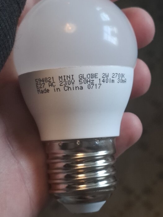 Närbild på en vit E27 glödlampa märkt "MINI GLOBE 2W 2700K, 230V". Lampan hålls i en hand, texten visar också tillverkad i Kina.