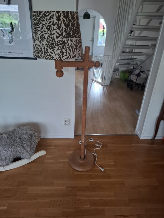 Golvlampa i trä med mönstrad lampskärm, synlig sladd och stickkontakt ligger på ett trägolv i ett rum med vita väggar och trappa i bakgrunden.
