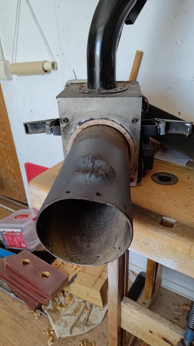 En trasig metallgavel och ett svart böjt rör på en arbetsbänk med träspån och verktyg i bakgrunden.