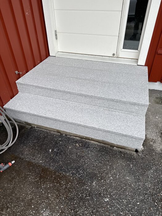 Grå trappa vid röd yttervägg och vit dörr, omgivande mark är delvis blöt.