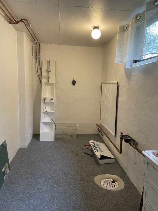 Källarutrymme med nedmonterad duschkabin, radiator, rörledningar, dräneringsbrunn och verktyg. Planer för ombyggnad till hobbyrum med skrivbord.