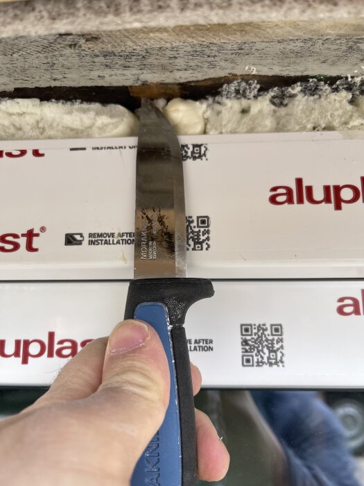 Hand håller en kniv vid glipa mellan fönsterram och vägg, isolering syns runt ramen med text "aluplast.