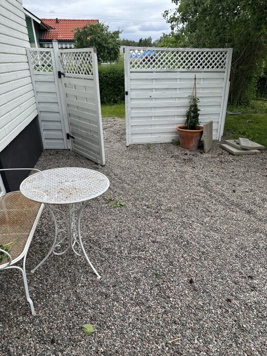 Grusgård med vitmålat trästaket, litet metallbord och bänk. Stenplattor och växt i kruka syns intill staketet.