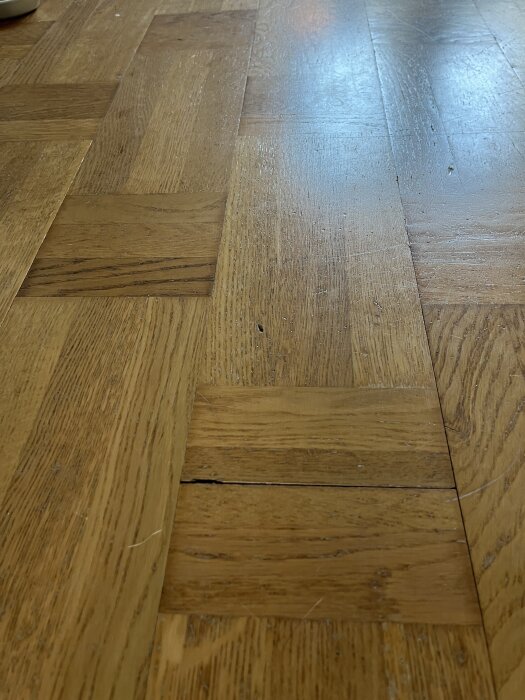 Parkettgolv med fiskbensmönster som visas i närbild, ytan ser lite sliten ut och behöver eventuellt behandling.